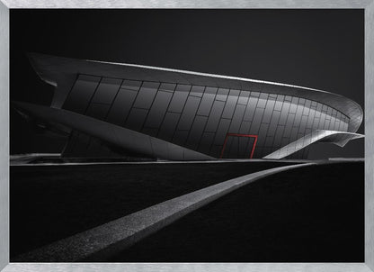 A dramatic black and white photograph of a futuristic, curved building with a dark, reflective facade. A single bright red door provides a striking pop of color. In the foreground, a light gray path curves across a dark landscape, leading the eye towards the structure. The entire image is presented within a silver-colored frame. Decor