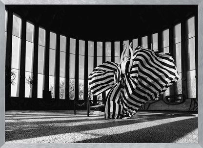 A dramatic black and white photograph of a woman in a voluminous, horizontally striped dress, captured mid-motion with the fabric swirling around her. She stands in a sunlit, circular room with tall, slatted windows that cast long shadows across the floor. Her arms are raised gracefully above her head, creating a striking and artistic pose. Artwork