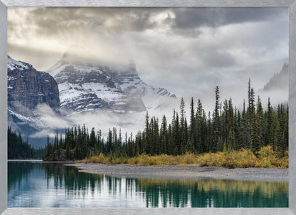 A tranquil mountain landscape featuring a still, teal lake reflecting a dense evergreen forest along its shore. In the background, majestic snow-covered peaks are shrouded in mist and dramatic gray clouds. Wall Art