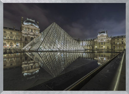 A dramatic night photograph of the illuminated Louvre Pyramid in Paris, its glass structure glowing against a dark, cloudy sky. The historic Louvre Palace is visible in the background, and the entire scene is perfectly reflected in the still water of a foreground pool. The image is presented within a silver frame. Artwork