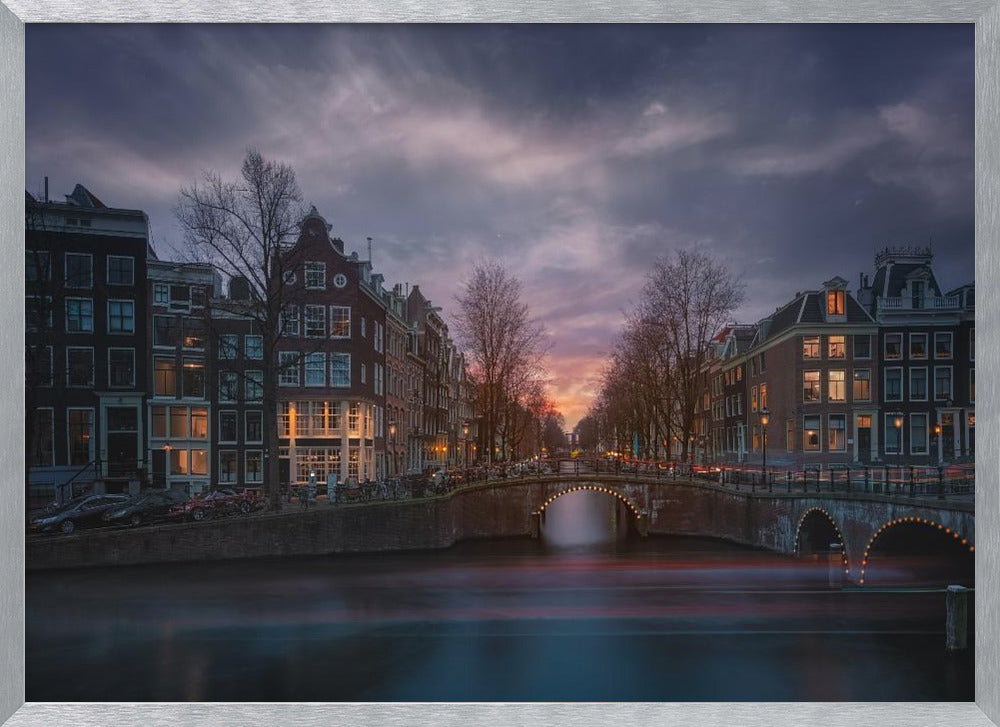 A dramatic long-exposure photograph of an Amsterdam canal at twilight. An arched bridge spans the water, lined by historic Dutch buildings with glowing windows. The sky is filled with dark, moody clouds above a fading orange and purple sunset, and red light trails from boats are blurred on the water's surface. Poster