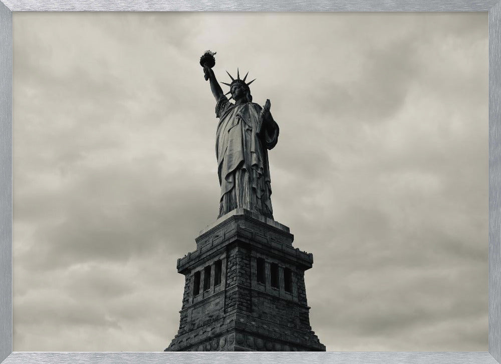 A low-angle, black and white photograph of the Statue of Liberty standing tall against a cloudy, dramatic sky, enclosed in a silver frame. Poster