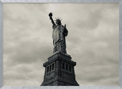 A low-angle, black and white photograph of the Statue of Liberty standing tall against a cloudy, dramatic sky, enclosed in a silver frame. Poster