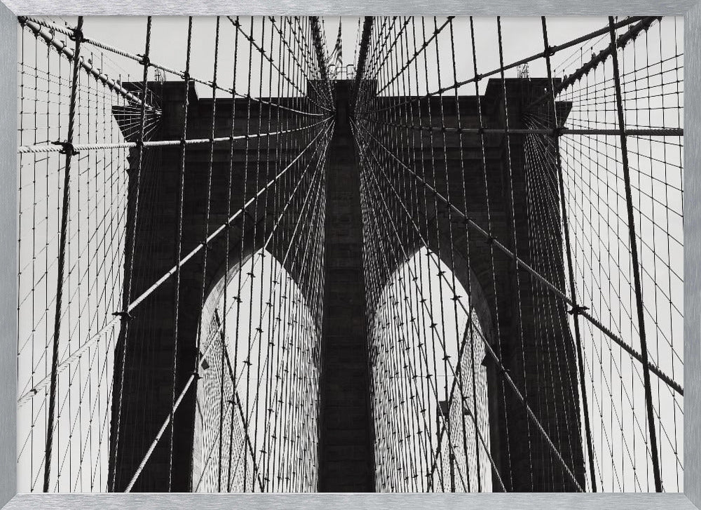 A low-angle, black and white photograph looking up at the iconic stone towers of the Brooklyn Bridge. The intricate web of suspension cables and vertical suspender ropes creates a symmetrical, geometric pattern against the light sky. The photo is enclosed in a silver frame. Print