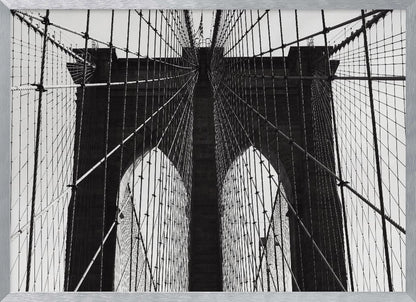 A low-angle, black and white photograph looking up at the iconic stone towers of the Brooklyn Bridge. The intricate web of suspension cables and vertical suspender ropes creates a symmetrical, geometric pattern against the light sky. The photo is enclosed in a silver frame. Print