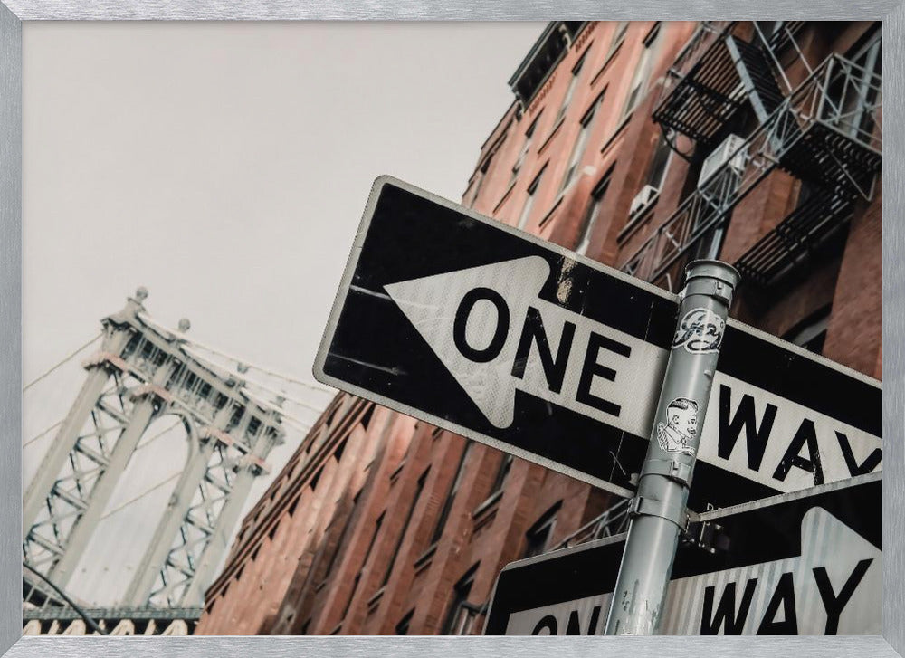 A low-angle shot of a black and white 'ONE WAY' street sign in the foreground, with a red brick building and the Manhattan Bridge visible in the background under an overcast sky, all enclosed in a silver frame. Artwork