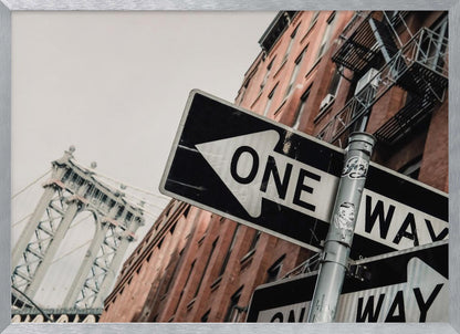 A low-angle shot of a black and white 'ONE WAY' street sign in the foreground, with a red brick building and the Manhattan Bridge visible in the background under an overcast sky, all enclosed in a silver frame. Artwork
