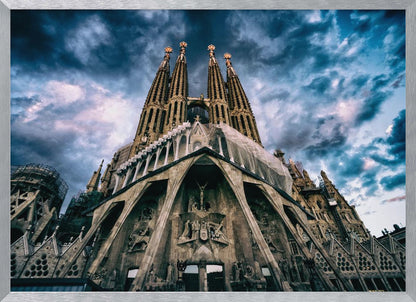 A dramatic, low-angle photograph of the Sagrada Familia in Barcelona, with its ornate stone facade and towering spires reaching up into a turbulent, dark blue and cloudy sky. The perspective emphasizes the grand scale and intricate detail of Gaudí's architecture. Print