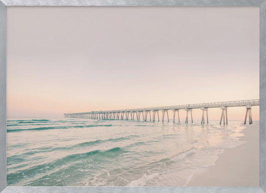 A tranquil beach scene with a long white pier extending into the turquoise ocean. The sky is a soft, pale pink, suggesting a serene sunrise or sunset, and gentle waves lap onto the sandy shore in the foreground. Poster