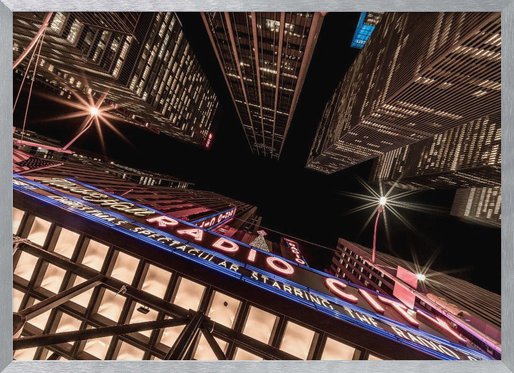 A dramatic low-angle shot looking up at the iconic Radio City Music Hall marquee at night, surrounded by towering, illuminated skyscrapers against a dark sky. Decor