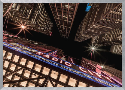 A dramatic low-angle shot looking up at the iconic Radio City Music Hall marquee at night, surrounded by towering, illuminated skyscrapers against a dark sky. Decor