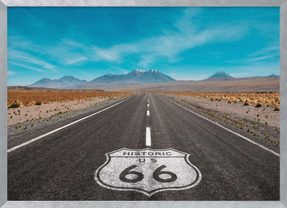 A framed photograph of the iconic Historic US Route 66 shield painted on a long, straight asphalt road that stretches through a desert landscape toward distant snow-capped mountains under a vibrant blue sky. Wall Art