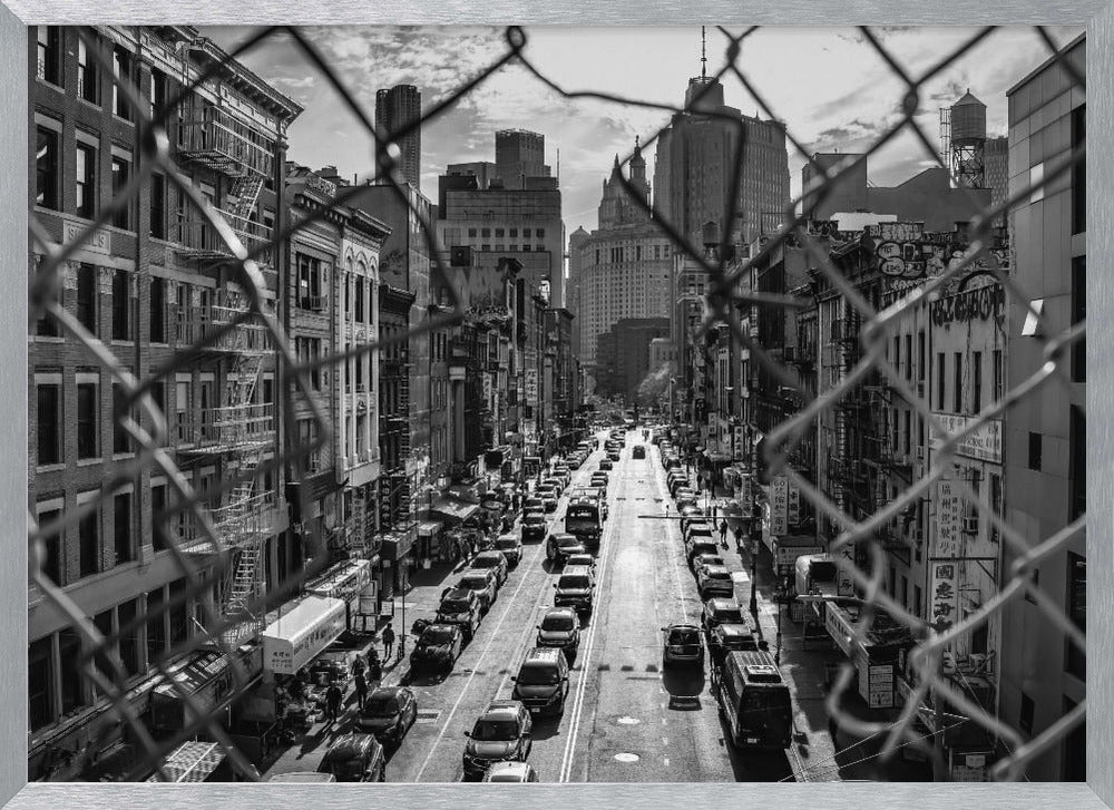 A high-angle, black and white photograph of a bustling New York City street viewed through a chain-link fence. The fence is in the foreground, framing the scene of traffic-filled streets, historic buildings with fire escapes, and skyscrapers in the distance under a cloudy sky. Wall Art