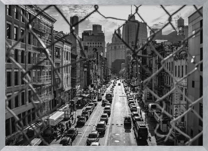 A high-angle, black and white photograph of a bustling New York City street viewed through a chain-link fence. The fence is in the foreground, framing the scene of traffic-filled streets, historic buildings with fire escapes, and skyscrapers in the distance under a cloudy sky. Wall Art