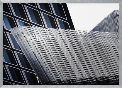 An abstract architectural photograph, framed in silver, showing a dynamic contrast between two modern building facades. In the background, a dark building features a grid of diamond-shaped windows with white frames, while a structure in the foreground creates a wave of fine, parallel white vertical lines against the sky and the other building. Print