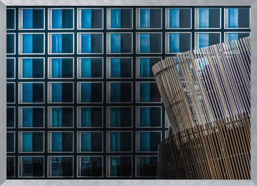 An architectural abstract photograph featuring a modern building facade with a grid of blue and black square windows, contrasted by a curved, slatted structure in the foreground, all enclosed in a silver frame. Wall Art