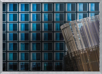 An architectural abstract photograph featuring a modern building facade with a grid of blue and black square windows, contrasted by a curved, slatted structure in the foreground, all enclosed in a silver frame. Wall Art