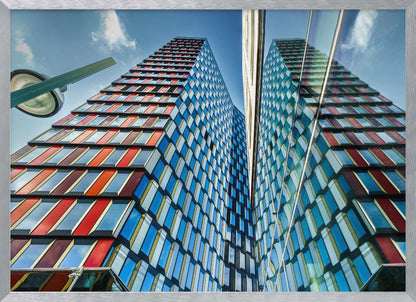 A low-angle shot of a modern skyscraper with a colorful facade of blue, red, and orange geometric panels, reflected in an adjacent glass building under a bright blue sky, all enclosed in a silver frame. Poster