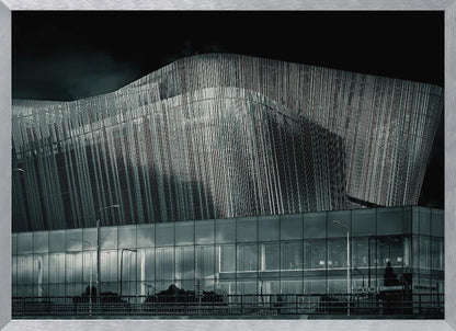 A dramatic black and white photograph of a modern building with a unique, wave-like facade made of vertical fins. The lower level is made of glass, reflecting the sky and streetlights. The entire image is presented in a silver frame. Artwork