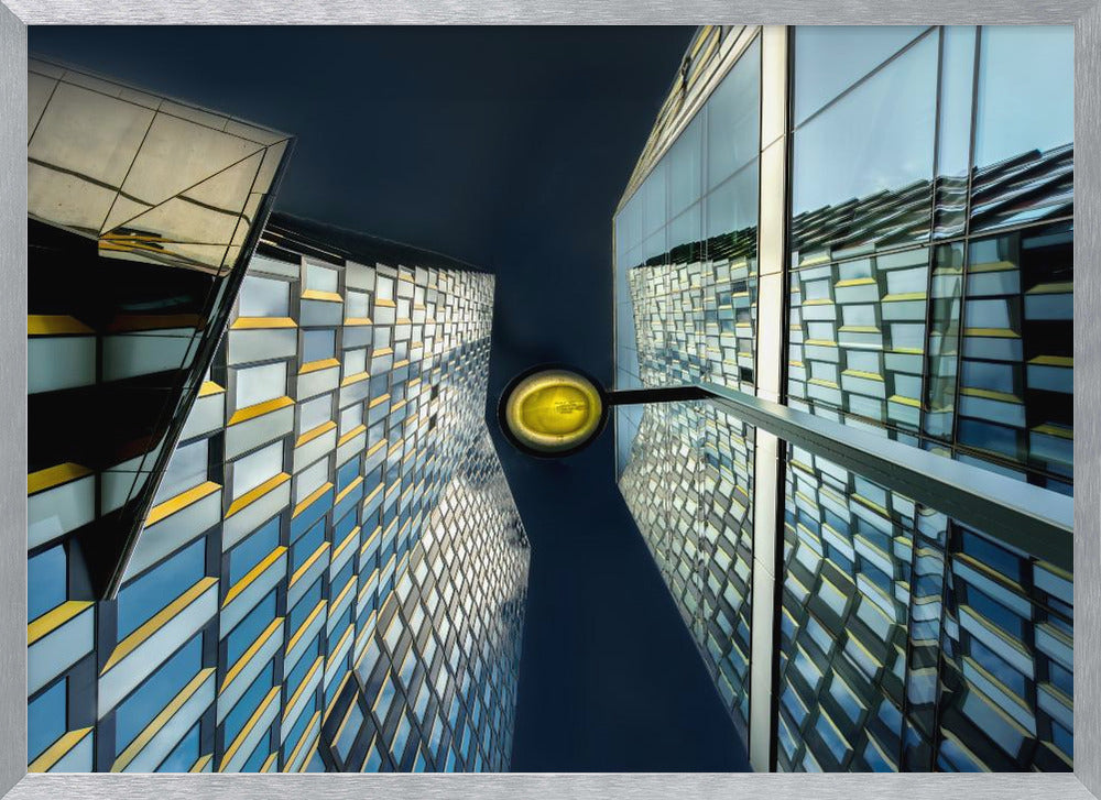 A worm's-eye view looking up between two modern glass skyscrapers with geometric blue, yellow, and silver panels, creating a symmetrical reflection against a dark blue sky, with a bright yellow streetlamp centered in the gap. Decor