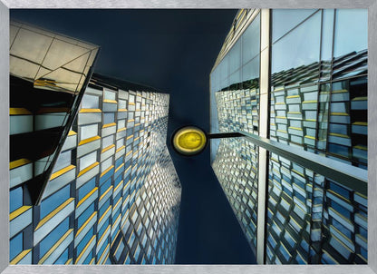 A worm's-eye view looking up between two modern glass skyscrapers with geometric blue, yellow, and silver panels, creating a symmetrical reflection against a dark blue sky, with a bright yellow streetlamp centered in the gap. Decor