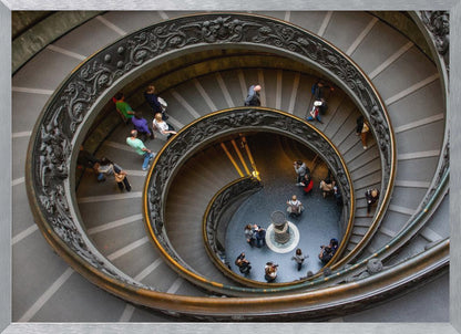 A framed, high-angle photograph looking down into the center of a grand spiral staircase. The gray stone steps spiral downwards, flanked by ornate, dark bronze railings. Numerous people are scattered along the stairs, appearing small from the overhead perspective, giving a sense of the structure's massive scale. Artwork