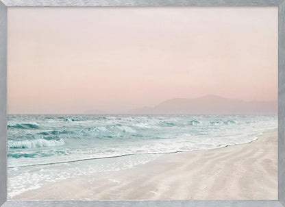 A serene coastal scene with gentle turquoise waves washing onto a sandy beach. The sky is a soft, hazy pink, and faint outlines of mountains are visible in the distant background. The artwork is presented in a silver frame. Print