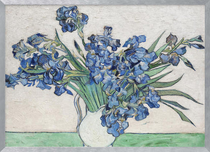 A framed print of Vincent van Gogh's famous painting 'Irises'. It features a large bouquet of vibrant blue iris flowers with long green leaves, arranged in a simple white pitcher. The background is a textured off-white wall, and the pitcher rests on a light green surface. The painting is in the post-impressionist style with thick, visible brushstrokes. Wall Art