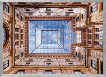 A worm's-eye view looking straight up from the center of an ornate, multi-story courtyard. The detailed walls create a symmetrical frame around a large glass skylight, which reflects a bright blue sky with fluffy white clouds. Print