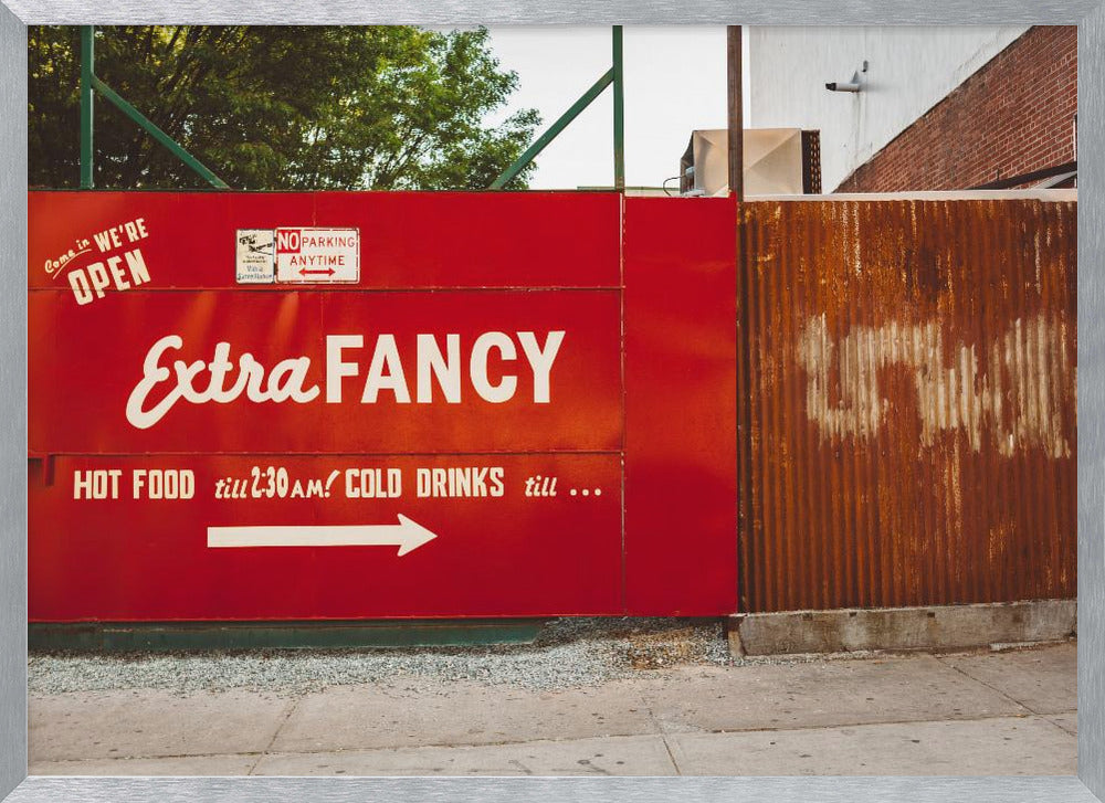 A framed photograph of a bright red outdoor sign for a restaurant named 'Extra Fancy'. The sign, painted with white retro-style text, advertises hot food and cold drinks and is situated next to a rusty, corrugated metal fence on a city sidewalk. Artwork