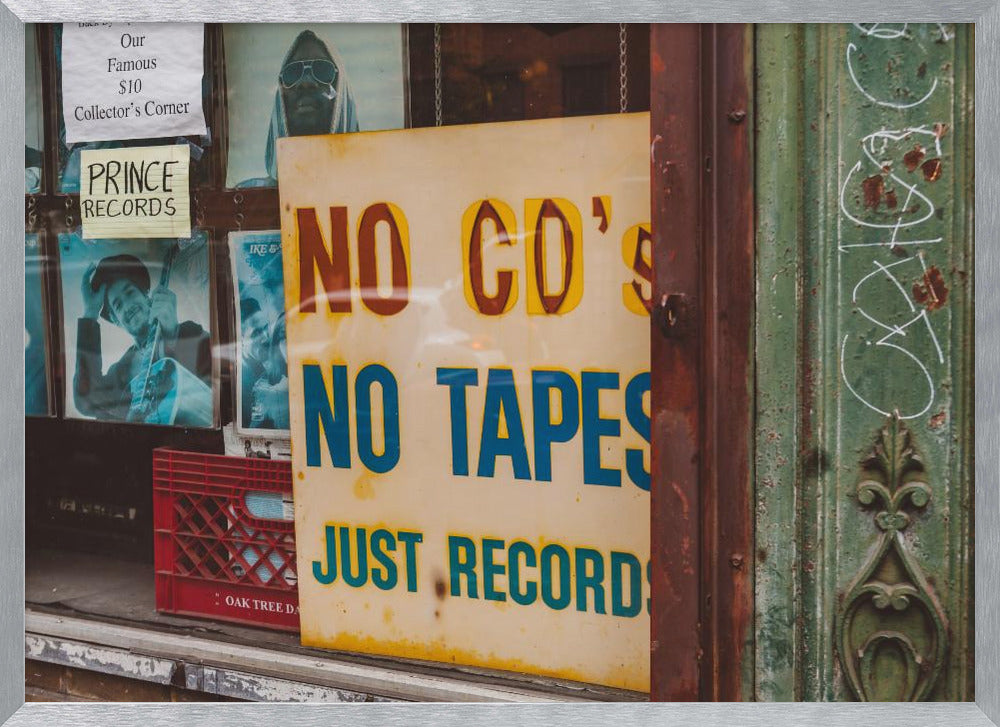 A weathered yellow sign in a record store window reads 'NO CD'S NO TAPES JUST RECORDS' in bold lettering. Behind the sign, vintage album covers and other posters are visible in the shop's display, next to a rustic green and brown door frame. Decor
