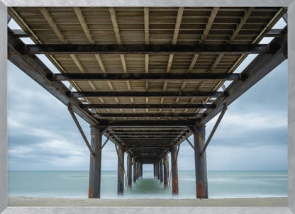 A symmetrical, low-angle photograph taken from underneath a long pier, looking out to sea. The rusty metal pilings and wooden planks of the pier create a receding perspective over the smooth, long-exposure water, all under a cloudy sky and presented within a silver frame. Poster