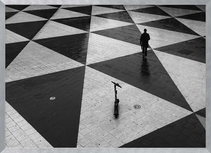 A high-angle, black and white photograph of a man in a coat and fedora walking away across a vast plaza with a geometric pattern of black and light gray triangles. An electric scooter stands alone in the foreground, creating a sense of solitude and contrast. The image is presented within a silver frame. Decor