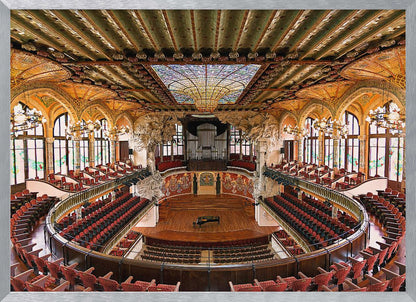 A high-angle, wide shot of the ornate and lavish interior of the Palau de la Música Catalana in Barcelona. The view captures the entire concert hall, with its curved balconies filled with red seats, a central wooden stage with a grand piano, and a spectacular, large stained-glass skylight in the ceiling. Poster