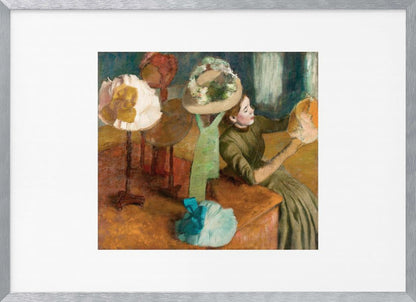 A framed print of Edgar Degas's Impressionist painting 'The Millinery Shop,' showing a woman in a green dress examining a hat in a shop filled with various fashionable hats on display stands. Print
