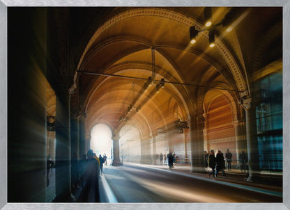 A long exposure photograph of a grand, vaulted hallway with brick arches. Dramatic beams of golden light cut through the space, creating horizontal streaks and a motion blur effect. Silhouettes of people walk through the atmospheric light and shadow. Wall Art
