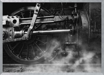 A dramatic, black and white close-up photograph of the wheels and driving rods of a steam locomotive. Puffs of steam rise from the tracks, partially obscuring the lower part of the machinery. Print