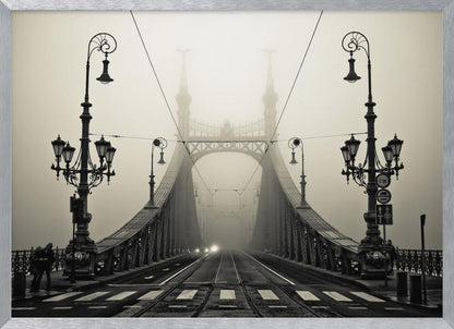 A framed, symmetrical black and white photograph of a large, ornate metal bridge disappearing into thick fog. Vintage lampposts stand on either side in the foreground, and the road and tram tracks lead the eye into the misty distance where faint car headlights glow. Print