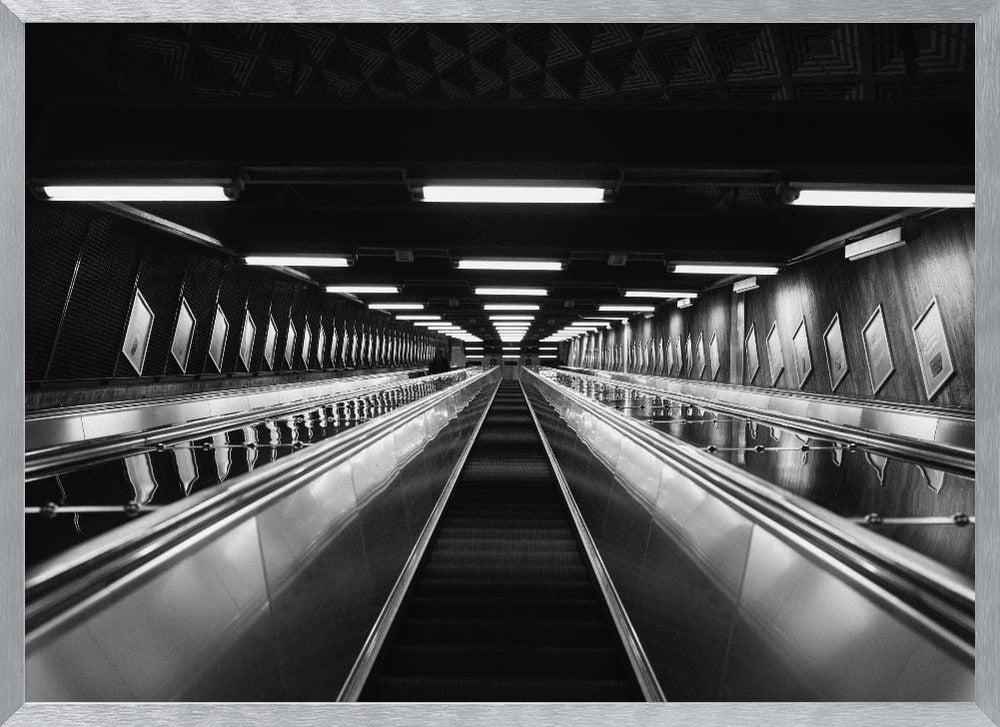 A framed, black and white, low-angle photograph looking up a long, steep, and empty escalator in a subway tunnel. The symmetrical composition uses the shiny metal handrails and rows of overhead lights as leading lines, creating a dramatic sense of depth and perspective. Print