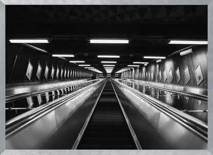 A framed, black and white, low-angle photograph looking up a long, steep, and empty escalator in a subway tunnel. The symmetrical composition uses the shiny metal handrails and rows of overhead lights as leading lines, creating a dramatic sense of depth and perspective. Print