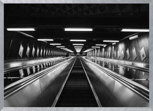 A framed, black and white, low-angle photograph looking up a long, steep, and empty escalator in a subway tunnel. The symmetrical composition uses the shiny metal handrails and rows of overhead lights as leading lines, creating a dramatic sense of depth and perspective. Print