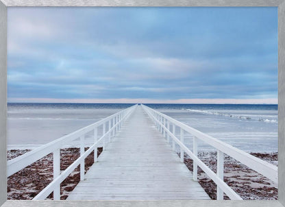 A framed photograph capturing a long, white wooden pier from a central perspective, leading the eye across a calm, icy sea towards the distant horizon under a cloudy blue sky. Print
