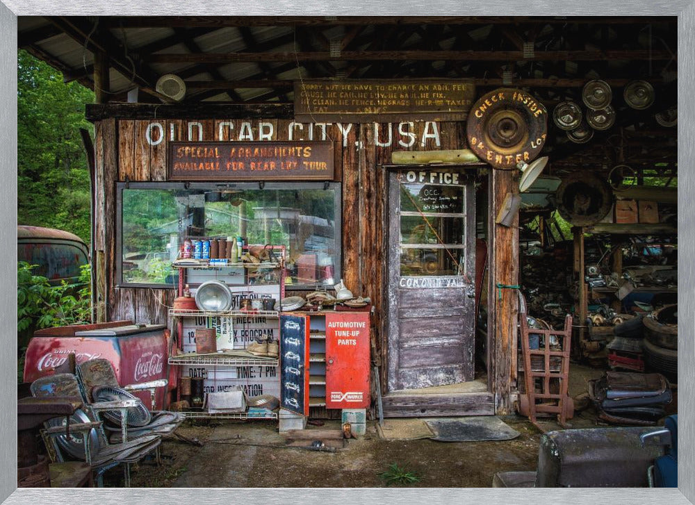 A framed photograph of the cluttered entrance to a rustic wooden building labeled 'Old Car City, USA'. The scene is filled with vintage memorabilia, including car parts, old signs, a red Coca-Cola cooler, and the weathered door to an office. Poster