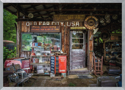 A framed photograph of the cluttered entrance to a rustic wooden building labeled 'Old Car City, USA'. The scene is filled with vintage memorabilia, including car parts, old signs, a red Coca-Cola cooler, and the weathered door to an office. Poster