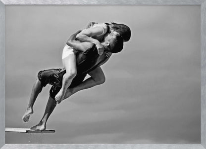 A framed, black and white photograph capturing a dynamic and romantic moment. A man is leaping off a diving board, carrying a woman who is wrapped around him in a passionate embrace, their faces close together. The background is a simple, plain gray, emphasizing their mid-air pose. Wall Art