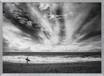 A dramatic black and white photograph of a lone surfer silhouetted against the sea, walking along a sandy beach. The sky is filled with spectacular, radiating clouds that stretch across the horizon, creating a powerful and moody atmosphere. The image is enclosed in a silver frame. Poster