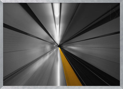 An abstract, long-exposure photograph from a first-person perspective inside a tunnel, creating a motion-blurred effect. The scene is mostly black and white, with lines radiating from a central vanishing point, but a single, bold yellow line runs down the center, adding a pop of selective color. The artwork is presented in a silver frame. Print