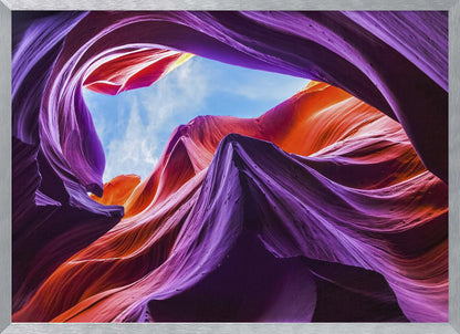 A low-angle photograph looking up from inside a vibrant slot canyon. The smooth, flowing sandstone walls are colored in rich shades of purple, red, and orange, curving around an opening that reveals a bright blue sky with wispy clouds. The image is presented within a silver-colored frame. Wall Art