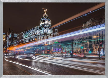 A dynamic long-exposure night photograph of the Metropolis Building in Madrid, with vibrant streaks of white, orange, and blue light from traffic swirling through the city streets. Artwork