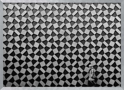 A black and white photograph of a lone person walking in front of a massive wall covered in a dizzying geometric pattern of tilted, textured gray quadrilaterals on a black background. The figure in the bottom right corner is dwarfed by the scale of the abstract architectural design. The image has a silver-gray frame. Artwork